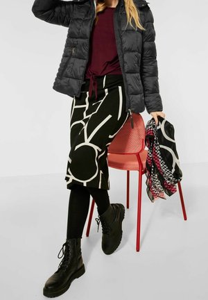 Sort pufferjakke, bordeaux top, knælang nederdel med hvide geometriske mønstre, sorte leggings og robuste sorte snørestøvler. Rød stol i nærheden.