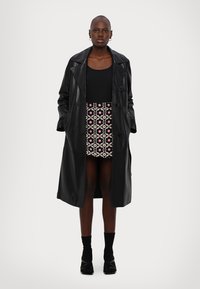 Schwarzer Ledermantel im Trenchcoat-Stil mit doppelter Knopfleiste, getragen über einem schwarzen Oberteil und einem gemusterten Minirock mit rosafarbenen und cremefarbenen Blumemotiven.