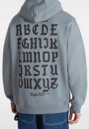 Personne portant un sweat à capuche gris clair avec des lettres gothiques noires alignées en rangées dans le dos, associé à un jean bleu.