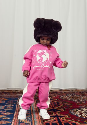Felpa rosa e pantaloni con strisce bianche ai lati, caratterizzati da un design a mappamondo. Accentuati da un cappello in peluche marrone scuro con pompon. Scarpa bianche.