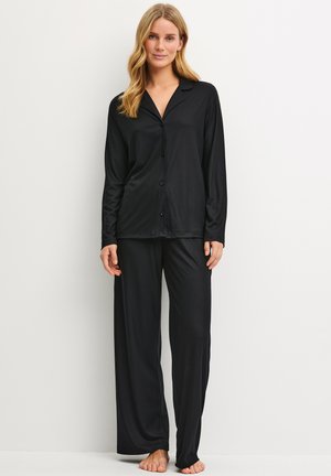 Femme debout pieds nus portant un haut noir ample à manches longues boutonné et un pantalon large assorti sur fond blanc.
