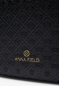 Superficie in pelle nera texture con motivi circolari in rilievo e un logo ed emblema dorato "Anna Field" centrato vicino al fondo.