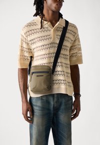 Polo tricotat de culoare bej deschis cu modele din dantelă. Geantă de umăr de culoare verde măslin cu accente negre și fermoar. Pantaloni din blugi de culoare bleumarin.