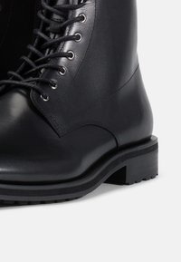 Botine din piele neagr�ă, cu șireturi, cu un finisaj neted, vârf rotund, talpă rezistentă și ochiuri pentru șireturi cu șireturi puternice pentru o potrivire sigură.