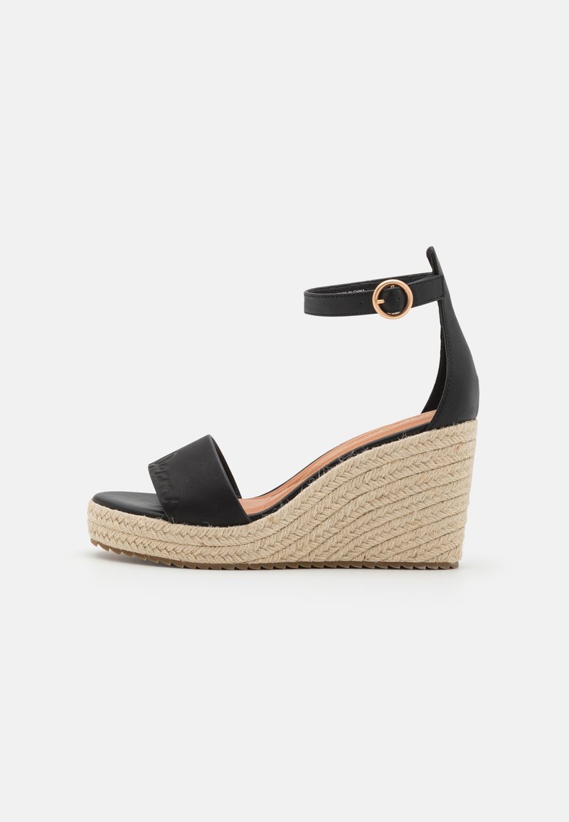 Svart kilehæl sandal med espadrille-design, som har en glatt overdel, ankelstropp med en sirkelbuckl og teksturert vevd såle.