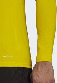 Närbild av en persons arm och midja som bär en gul långärmad tröja med "AEROREADY"-text ovanför svarta byxor.