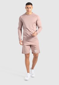 Helles rosa, langärmeliges Sportoberteil kombiniert mit passenden Shorts. Auffälliges weißes Logo auf den Shorts. Getragen mit weißen Turnschuhen.