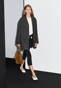 Przesadnie duża szara marynarka, biała koszula, czarne legginsy i białe baleriny. Trzyma delikatną brązową torebkę z zamszu. Prosta srebrna naszyjnik.
