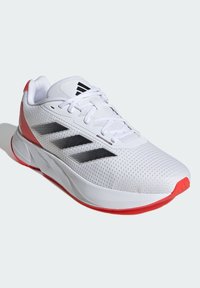 Zapatilla deportiva blanca con parte superior de malla, tres rayas negras y un acento rojo en el talón. Presenta una suela acolchada y un diseño de cordones.