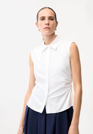 Kvinde med trukket hår tilbage, iført hvid ærmeløs skjorte med krave og marineblå plisseret nederdel, stående foran en ensfarvet hvid baggrund.
