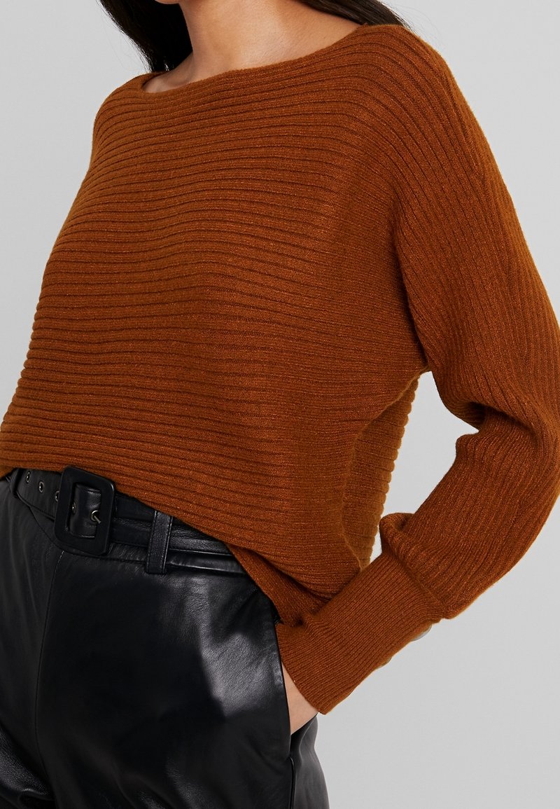 Brauner gerippter Pullover mit weitem Ausschnitt und langen Ärmeln, kombiniert mit schwarzen, hochtaillierten Lederhosen mit einem Gürtel.