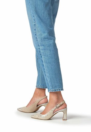 Piernas con pantalones vaqueros azul claro combinados con tacones altos slingback beige con un sutil estampado floral y detalle de hebilla.