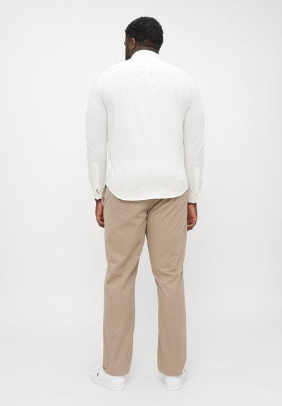 Chemise blanche à manches longues avec des poignets à boutons, associée à un pantalon beige et des chaussures blanches, photographiée de dos devant un fond neutre.