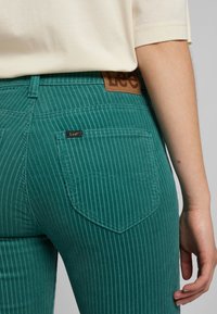 Teal vertikálně pruhované džíny z denimu s koženou etiketou a jednou zadní kapsou. Materiál je texturovaný, s viditelnými detaily šití.