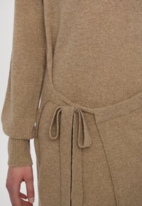 Pull en maille beige avec une finition texturée, manches ajustées et un nœud à la taille pour un style ajustable.