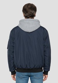 Marineblaue Bomberjacke mit grauer Kapuze. Verfügt über elastische Bündchen und Saum, gesteppte Textur und Seitentaschen. Sichtbare Nähte.