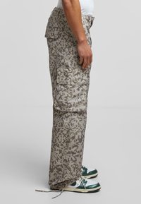 Cargo bukser i et gråt camouflage-mønster med flere sidelommer, snøre i kanten og let, glat materiale. Stylet med grønne og hvide sneakers.