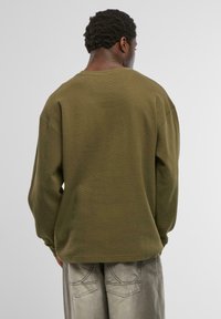 Olivzaļš džemperis ar garām piedurknēm, izgatavots no teksturēta, ribota materiāla. Tam ir brīvs piegriezums un nolaistas plecu daļas, skatīts no aizmugures.