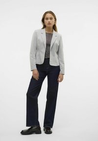 Blazer gris claro con cuello de solapa, llevado sobre una blusa gris oscuro. Combinado con jeans anchos de color azul oscuro y zapatos de vestir negros. Textura de tela suave.