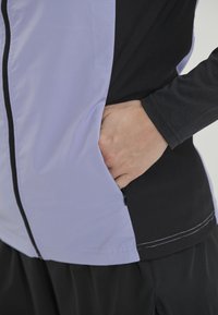 Lichte sportjack met een lavendelkleurig voorkant en een zwart lichaam, met een ritszak aan de zijkant en contrasterende stiksels.