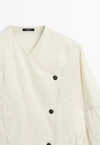 Off-white wrap-style blouse with three black buttons on the front and a V-neck collar, made from lightweight textured fabric.
