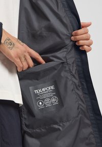 Schwarze Texapore Ecosphere Pro-Jacke von innen mit einem Etikett, das die wasserdichten, atmungsaktiven und winddichten Eigenschaften beschreibt. Seitentasche sichtbar.