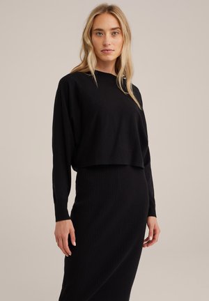 Pullover corto nero con maniche lunghe, realizzato in tessuto morbido; abbinato a una gonna midi nera a coste, caratterizzata da una leggera variazione di texture.