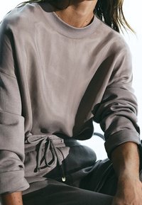 Personne portant un sweat-shirt ample mauve aux manches retroussées et un pantalon à cordon assorti, assise dans une lumière naturelle douce.