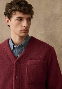 Burgunderfarbener Fleecepullover mit V-Ausschnitt, zwei Knöpfen und einer Vordertasche, über einem hellblauen Jeanshemd getragen. Texturiertes und warmes Design.