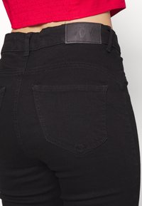 Vaqueros negros con una textura suave, cintura de altura media y sutiles bolsillos traseros. Presenta un parche de marca de cuero negro en la parte trasera.