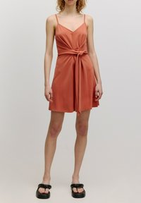 Rostfarbenes Kleid mit tailliertem Oberteil und Bindedetail in der Taille. Verfügt über dünne Träger und einen ausgestellten Rock. Wird mit schwarzen Sandalen getragen.