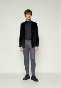 Blazer en velours noir sur une chemise noire, associé à un pantalon slim gris et des chaussures noires à lacets, sur un fond neutre.