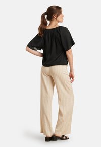 Schwarze Bluse mit kurzen Ärmeln und gerafftem Rücken, kombiniert mit beigen gestreiften weit geschnittenen Hosen und schwarzen Sandalen mit Blockabsatz.