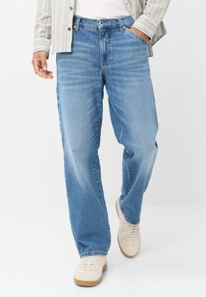 Mand iført lyseblå jeans med lige ben, hvide sneakers, en hvid t-shirt og en beige stribet skjorte med knapper, med den ene hånd i lommen.