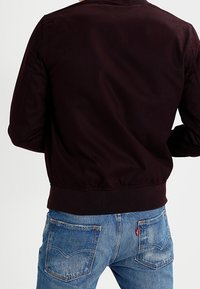 Persoon in een donkerbruine jas en blauwe spijkerbroek, gezien vanaf de achterkant van de taille tot de schouders.