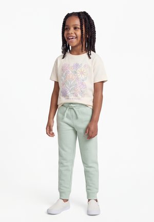 Enfant souriant aux cheveux tressés portant un t-shirt crème à imprimé floral, un pantalon de jogging vert menthe et des chaussures blanches sans lacets, debout sur un fond blanc.