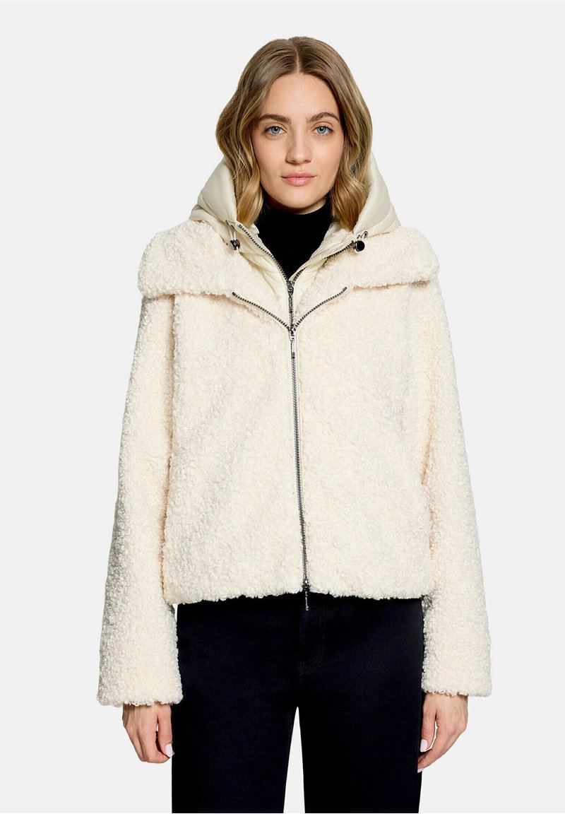 Femme aux cheveux châtain clair portant une veste zippée texturée couleur crème sur un top noir, debout devant un fond uni.