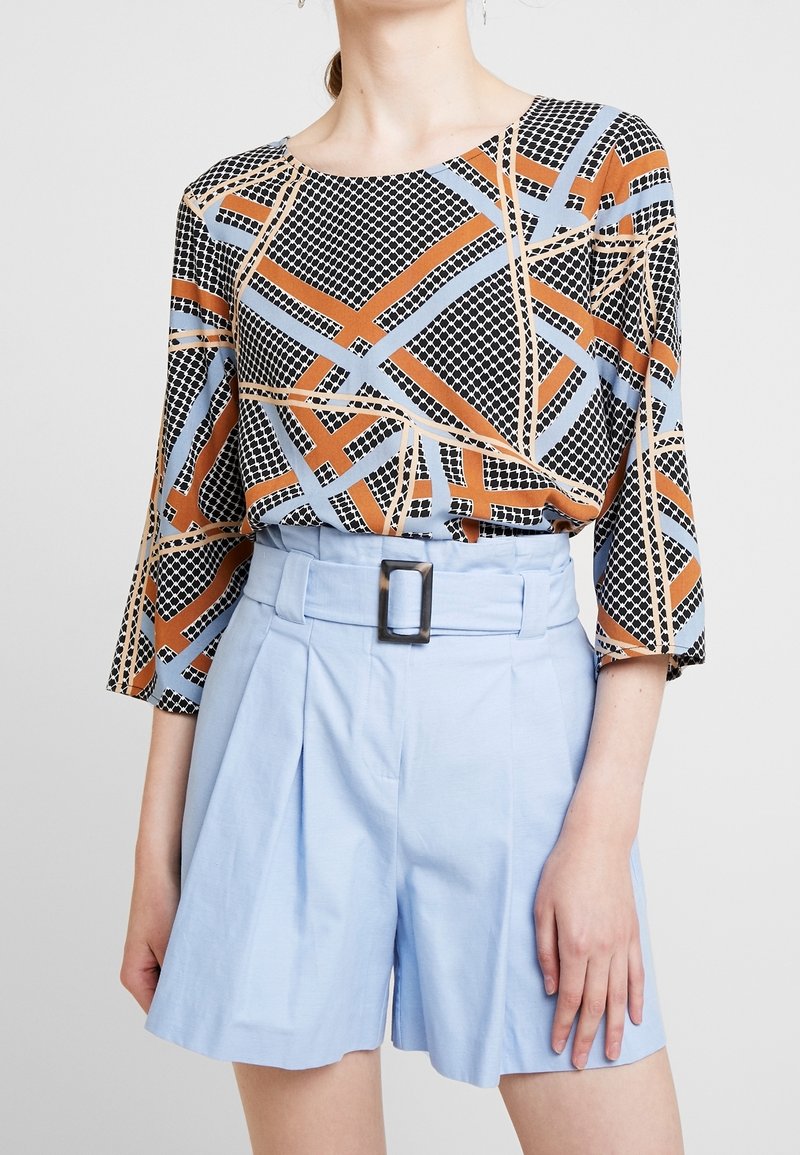 Multifarvet bluse med geometriske mønstre i sort, orange og blå, parret med lyseblå shorts med bælte og plisseringer.