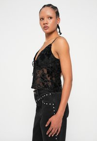 Melnas mežģīņu kamzolis ar ziedu rakstiem un dziļu V veida kakla izgriezumu. Caurspīdīgs audums, plānas siksniņas, apvienots ar augstās jostasvietas melnajām džinsām, kas rotātas ar uzrakstiem.