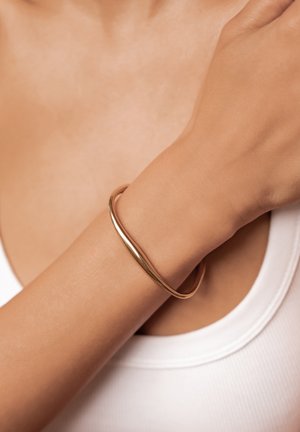 Woman wearing a smooth, shiny gold twisted bangle bracelet on her wrist, against a white tank top and neutral skin tone background.