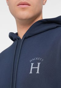 Μπλέ φούτερ από μαλακό ύφασμα, με λογότυπο μπροστά που λέει "HACKETT" πάνω από ένα έντονο γράμμα "H". Ανεμολόγος σχέδιο με κουκούλα που έχει κορδόνι.