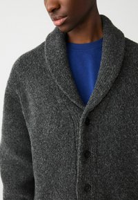 Gray knitted cardigan with a shawl collar, featuring a textured finish and four black buttons, worn over a blue shirt.