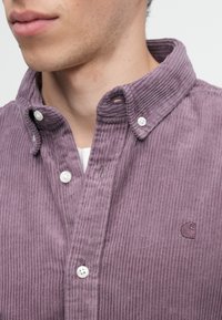 Fialová kordové košeľa s golierom na gombíky, so bielymi gombíkmi a vyšitým logom na hrudi. Textúrovaná tkanina a rebrovaný dizajn.