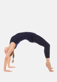 Fekete sporttop és leggings, női hídpozícióban, kezén egyensúlyozva, a rugalmasságát bemutatva egy fehér háttér előtt.
