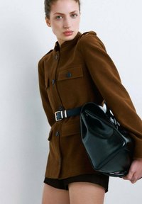 Veste en mélange de laine marron avec deux poches avant, boutons noirs, et une ceinture ; portée avec un short foncé et un sac à main en cuir noir.