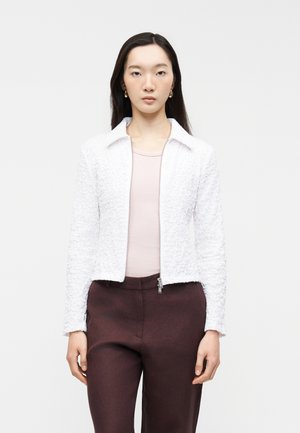 Γυναίκα με ίσια σκούρα μαλλιά φορά χρυσούς κρίκους σκουλαρίκια, λευκό υφασμάτινο σακάκι με φερμουάρ, ροζ ανοιχτό μπλουζάκι και σκούρο καφέ παντελόνι, στέκεται μπροστά σε λευκό φόντο.