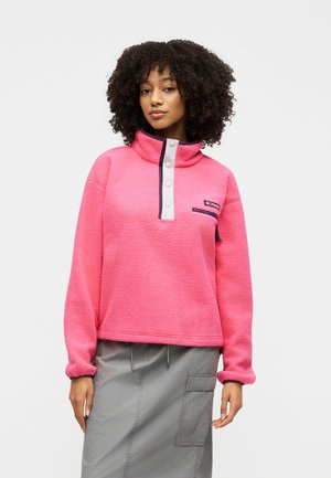Mujer con cabello rizado que lleva un jersey polar rosa con botones a presión y una falda gris con un bolsillo tipo cargo sobre un fondo liso.