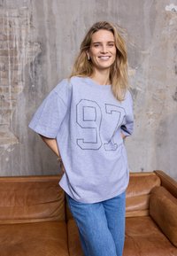 Halli üle suurusega T-särk, millel on number "97" kontuurse disainiga. Ühendatud helesiniste teksadega, taustal tekstuuriga pind.