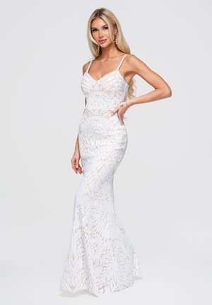 Femme aux longs cheveux blonds portant une robe blanche cintrée sans manches avec un motif, posant avec une main sur la hanche sur un fond uni.