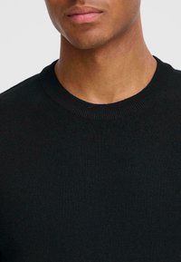 Pulover negru tricota cu guler rotund, având o textură netedă și manșete cu nervuri. Vedere de aproape fără modele sau accesorii vizibile.